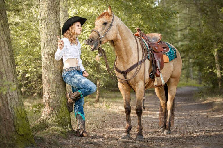 Mädchen im Westernoutfit steht mit einem hellen Pferd auf einem Waldweg