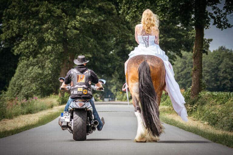 Braut reitet auf einem Pferd neben einem Motorradfahrer über eine von Bäumen gesäumte Landstraße